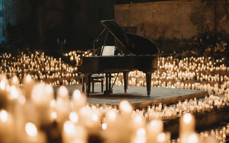 Zdjęcie przedstawia fortepian, który jest otoczony zapalonymi świecami