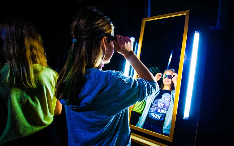 Zdjęcie przedstawia dziewczynkę przymierzającą okulary w lustrze na wystawie