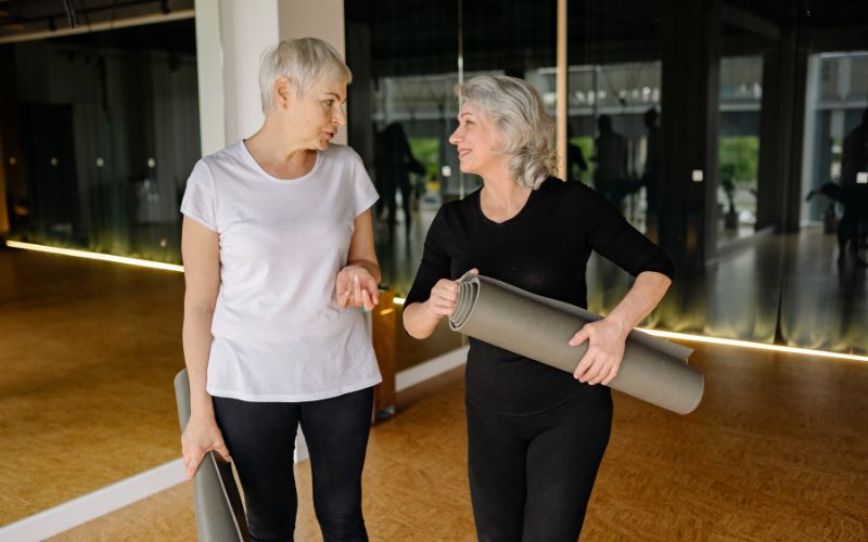 Zdjęcie przedstawia dwie kobiety na emeryturze, które trzymają w rękach maty ćwiczące i są na sali treningowej