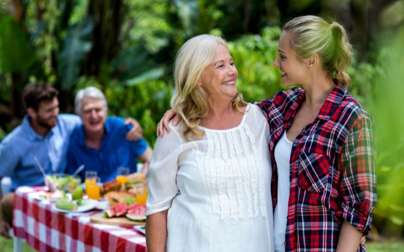 Zdjęcie na pierwszym planie przedstawia teściową z synową, za nimi znajduje się stół przy którym siedzą mężowie