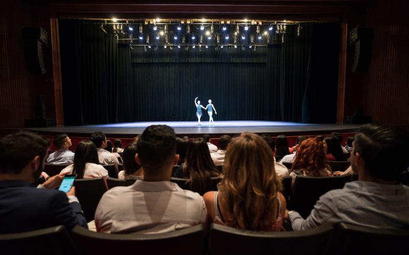 Zdjęcie wykonane na sali teatralnej zza publiczności. Na scenie dwie osoby tańczą
